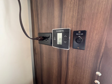 RV rental interior controls: Dometic thermostat, plugged-in outlet, and bathroom light switch on a wood panel.