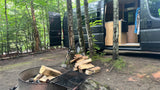 RV rental adventure: Camper van parked at a wooded campsite with a fire pit and stacked firewood, ready for use.