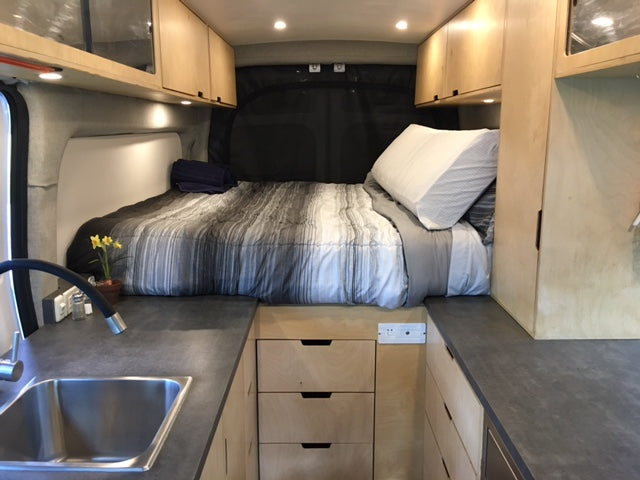 This rv rental features a cozy bed, kitchen sink, and wood cabinets, offering comfort in a compact van interior.