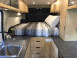 This rv rental features a cozy bed, kitchen sink, and wood cabinets, offering comfort in a compact van interior.