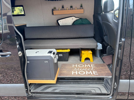 View inside an RV rental camper van with a bench bed, portable fridge, storage, & 'Home Sweet Home' mat. Ready for adventure!