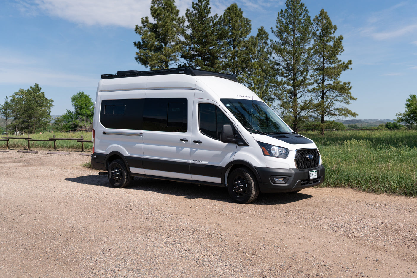 White adventure camper van, ideal for RV rental and outdoor road trips, parked on a gravel path with trees and blue sky.