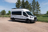 White adventure camper van, ideal for RV rental and outdoor road trips, parked on a gravel path with trees and blue sky.