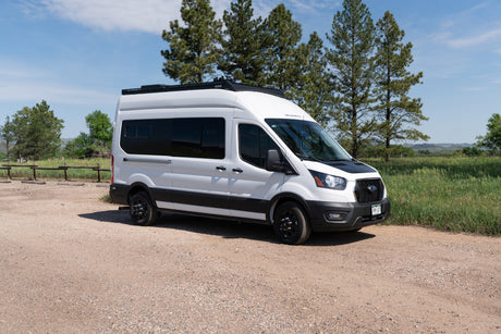 White adventure camper van, ideal for RV rental and outdoor road trips, parked on a gravel path with trees and blue sky.