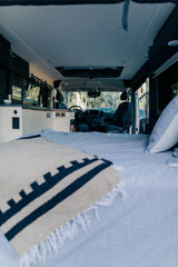 Inside a cozy RV rental: a comfortable bed with a patterned blanket leads to a built-in kitchenette and the driver's area beyond.