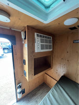 Compact RV rental interior: wooden walls, window AC unit, skylight, and a small bed create a functional living space.