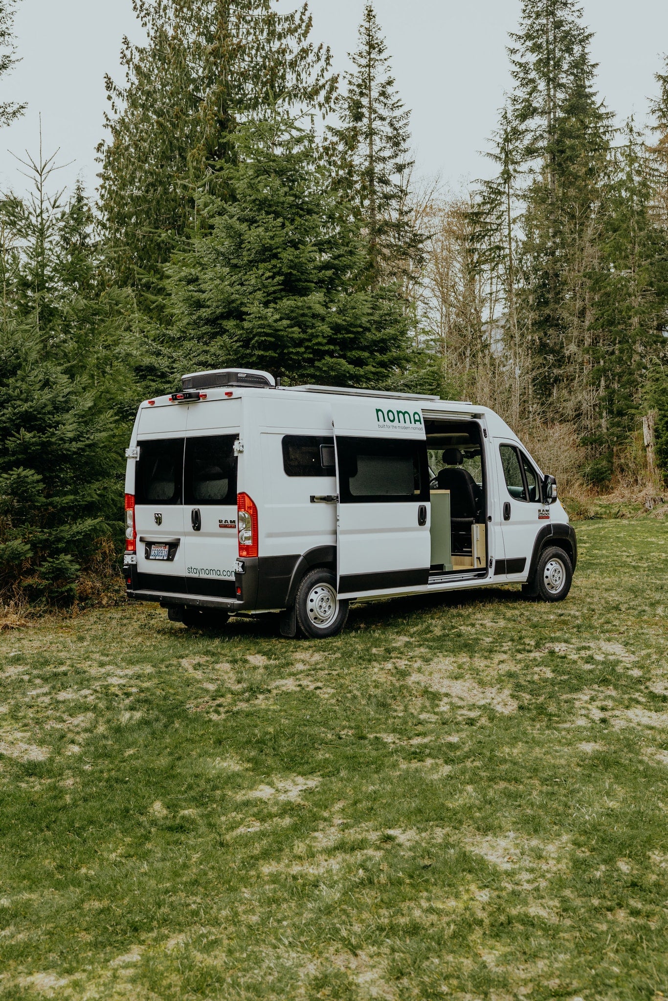 Noma campervan RV rental, a white Ram ProMaster van, parked on grass with its side door open, surrounded by trees. — photo 18