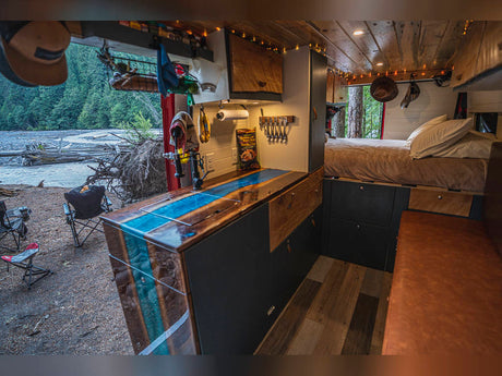 This RV rental shows a custom camper van's interior: kitchen with blue epoxy counter, bed, and a scenic river view outside.