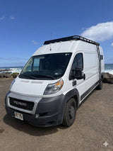 A white Ram ProMaster rv rental van, equipped with a roof rack, parked oceanfront under a bright blue sky for van life adventures.