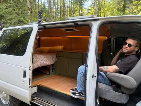 A man sits in his converted RV rental van, showcasing its custom bed, cabinets, and lighting for outdoor adventures.