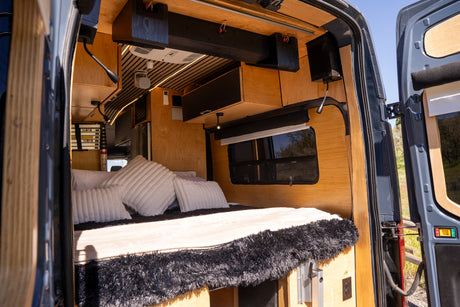 Cozy rv rental interior: A custom-built camper van with a bed, wooden walls, and modern amenities, viewed from the open back doors.
