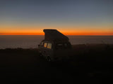 Experience stunning ocean sunset views with an RV rental. A silhouetted camper van with a pop-top roof overlooks the vibrant horizon.