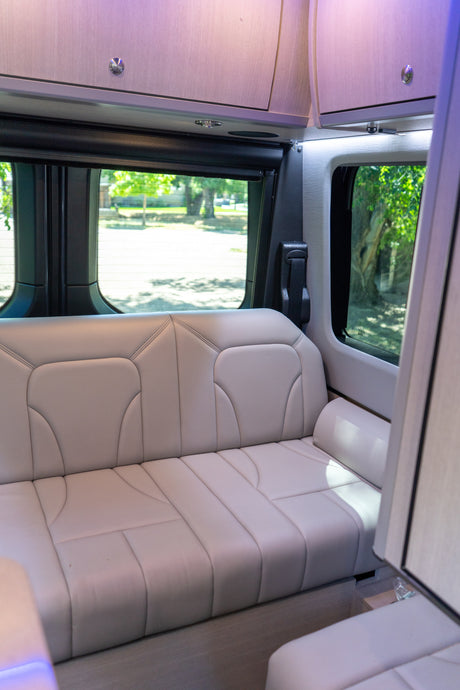 Modern RV rental interior with light grey bench seat, overhead cabinets, and windows offering a view outside.