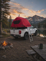 Adventure awaits with this RV rental alternative: a white truck featuring a red rooftop tent, campfire, and scenic mountain views.