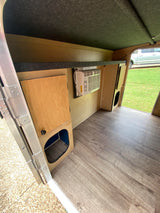 RV rental interior: light wood floor, wall-mounted AC, wooden cabinet, and storage cubby. Gray carpeted ceiling.