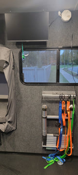 RV rental interior view: Mounted TV, rainy window, utility ladder, and tie-down straps stored on carpeted walls.