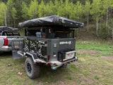 Adventure-ready overland trailer **RV rental** for camping, equipped with a rooftop tent, parked in a lush forest.