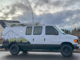 White custom camper van with mountain graphics & roof rack, perfect for rv rental adventures. Parked outdoors on wet asphalt.