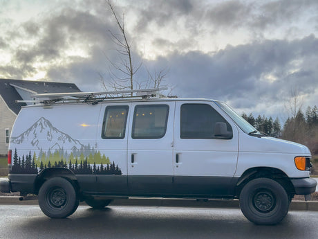 White custom camper van with mountain graphics & roof rack, perfect for rv rental adventures. Parked outdoors on wet asphalt.