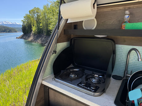 RV rental outdoor kitchen with stove, sink, and paper towels, overlooking a stunning lake, green trees, and snow-capped mountains.