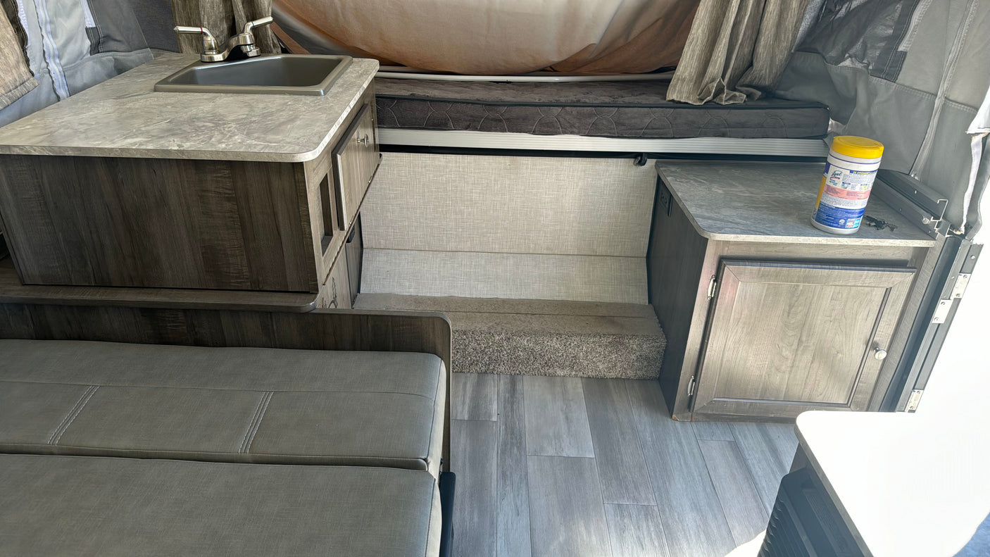 Clean rv rental interior showcasing a compact kitchen with sink, dark wood cabinets, grey bench seating, and vinyl flooring. Bed area visible.