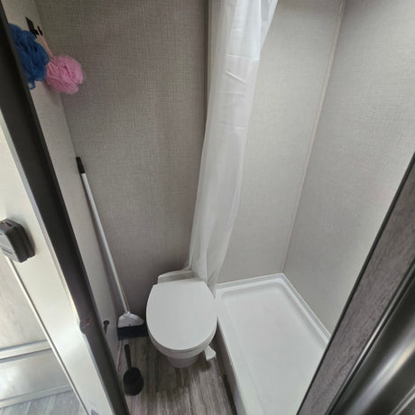 This RV rental's compact bathroom features a toilet, shower stall with curtain, bath poufs, and cleaning tools.