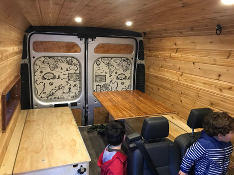 Spacious, wood-paneled RV rental interior features a folding table, bed platform, custom map doors, and seating, ideal for family travel.