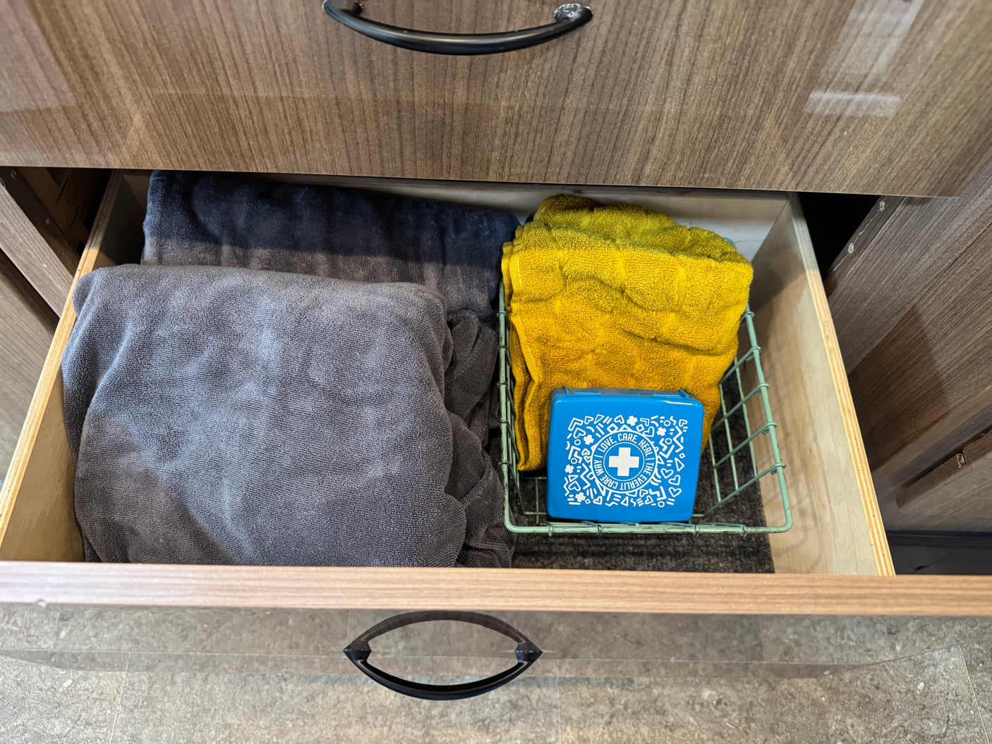 Open RV rental drawer showing gray/yellow towels & a blue first aid kit in a wire basket.