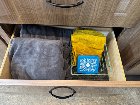 Open RV rental drawer showing gray/yellow towels & a blue first aid kit in a wire basket.