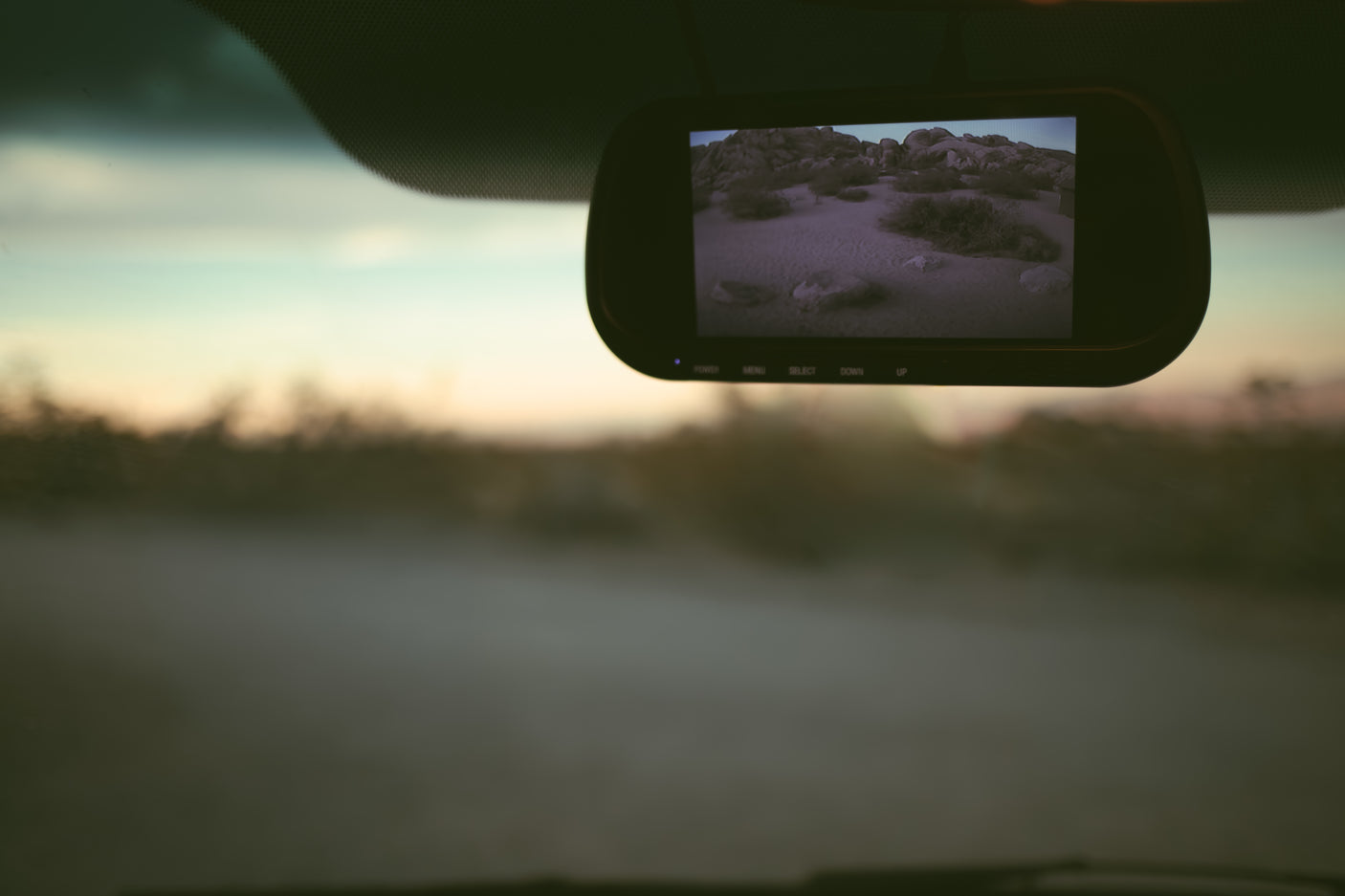 Inside an RV rental, the rearview mirror screen displays a rocky desert scene, contrasting with the blurred, dusty road outside at sunset.