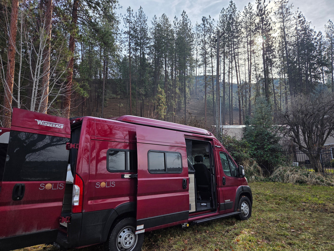 Red Winnebago Solis RV rental parked in a forest clearing with its side and rear doors open.