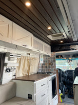 Inside this cozy RV rental: a modern camper van interior with a well-equipped kitchenette, stylish dark wood ceiling, and white cabinetry.