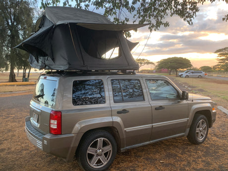 This RV rental, a Jeep Patriot SUV with a deployed rooftop tent, offers outdoor adventure at sunset.