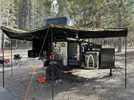 An off-road camper trailer at a wooded campsite, with awning, pull-out kitchen, and bed. Perfect for adventure RV rental!