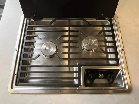 Compact Dometic 2-burner gas cooktop in an RV rental kitchen, with black grates and control knobs.