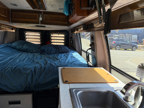 Cozy RV rental interior with blue bed, kitchenette, and sink. Perfect for travel adventures, viewing street outside.