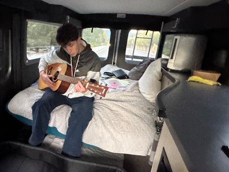 Young man playing guitar on the cozy bed of an RV rental, enjoying music during his journey.