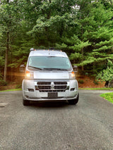 Silver RV rental van parked on an asphalt driveway, illuminated headlights against a lush green forest background.