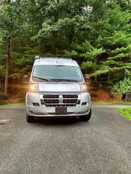 Silver RV rental van parked on an asphalt driveway, illuminated headlights against a lush green forest background.