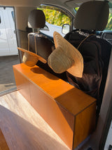 RV rental interior: custom wooden storage and floor, two straw hats hanging from front seats. Sunny view with palm trees.