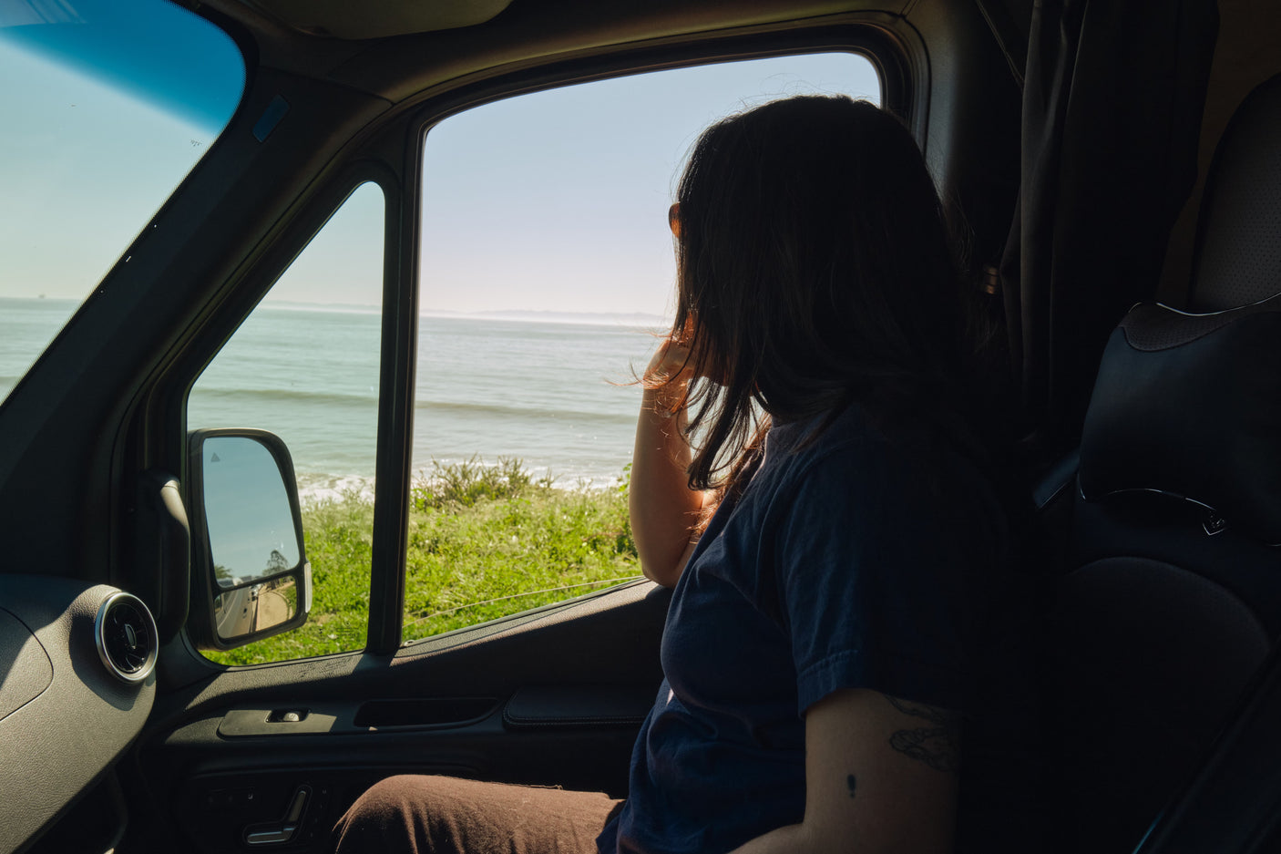 Ocean view from an rv rental. A traveler gazes out a van window at the bright ocean waves and green coastline on a sunny day.