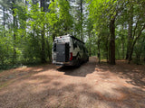 White camper van RV rental parked on a gravel path in a sunny forest, ready for adventure.