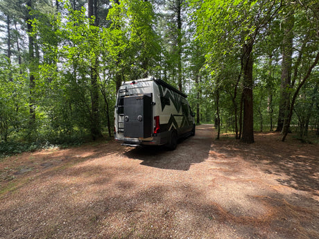 White camper van RV rental parked on a gravel path in a sunny forest, ready for adventure.