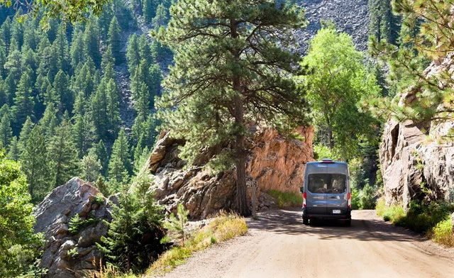 Gray RV rental van on a dirt mountain road surrounded by tall pines and rocky cliffs.