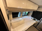 A cozy RV rental interior featuring a neatly made bed, spacious overhead cabinets, and windows, offering comfort and convenience.