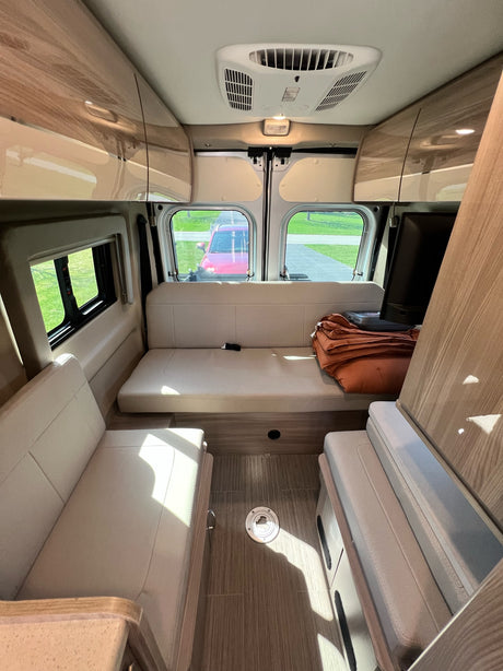 Interior of a modern RV rental, featuring light-colored benches converting to a bed, overhead cabinets, and a view out the back.