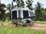 A grey pop-up camper trailer, ideal for an RV rental, is set up in a serene forest campsite.
