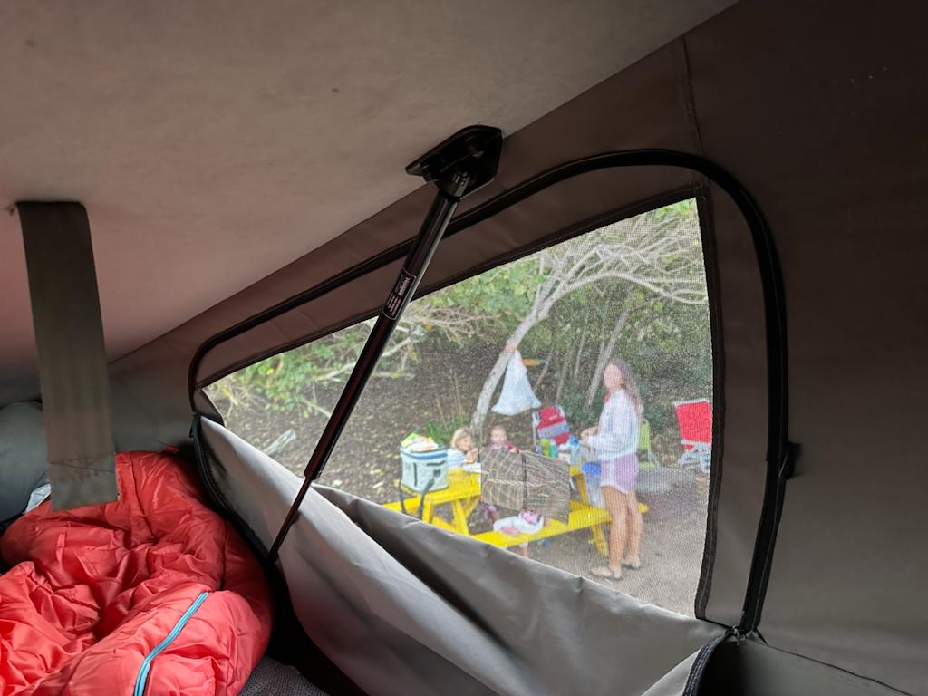 Inside an RV rental: a window with a gas strut opens to a screened view of a family enjoying a sunny campsite with a yellow table.