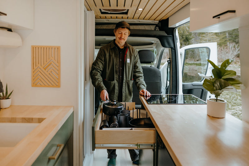 A man in an RV rental kitchen pulls cookware from a drawer, ready to cook in the modern camper van amidst nature. — photo 7