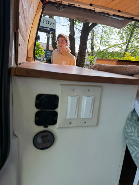 Interior view of an RV rental van conversion, showing a man outside, electrical panel with outlets & switches.